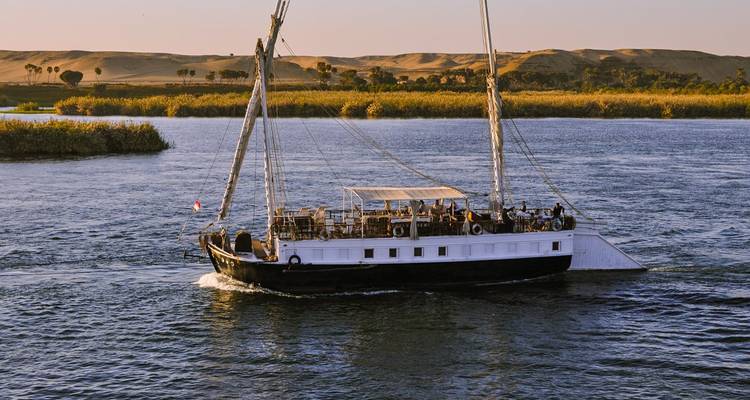 Gran velero navegando por un río con personas a bordo y paisaje desértico al fondo.