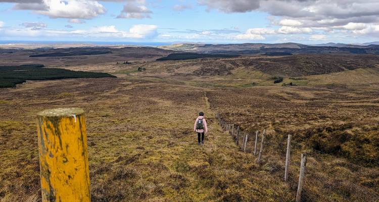 Solo hiker traverses open moorland with vast rolling countryside beyond