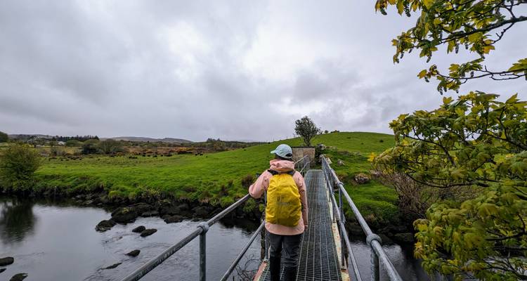 Hiker wearing a yellow backpack crosses a narrow footbridge over a peaceful Irish stream under overcast skies.