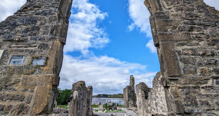 Stone ruins framing a view toward a small cemetery and blue sky beside an Irish lake.