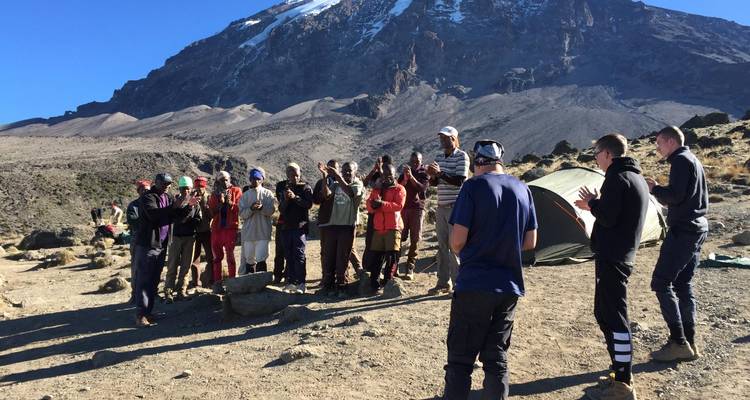 Klimmers and dragers verzamelen zich in een halve cirkel bij een rotsachtig kamp onder de dreigende top van de Kilimanjaro.