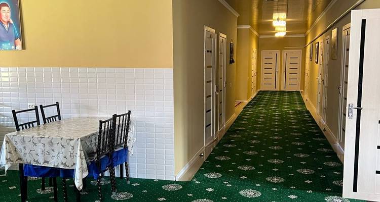 Couloir d'auberge simple avec tapis vert, plusieurs portes et un petit coin repas avec table et chaises.