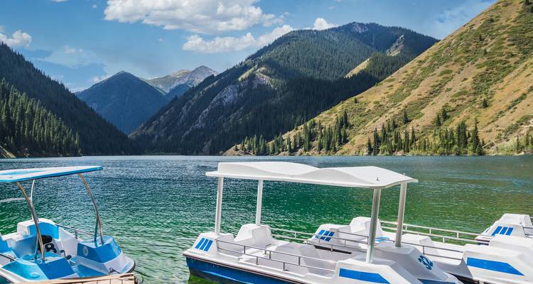 Des pédalos amarrés sur un lac alpin cristallin entouré de collines densément boisées de conifères et de pics montagneux.