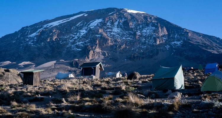 Tiendas y cabañas en un campamento de altura bajo la cumbre nevada y escarpada del Kilimanjaro en la luz del amanecer