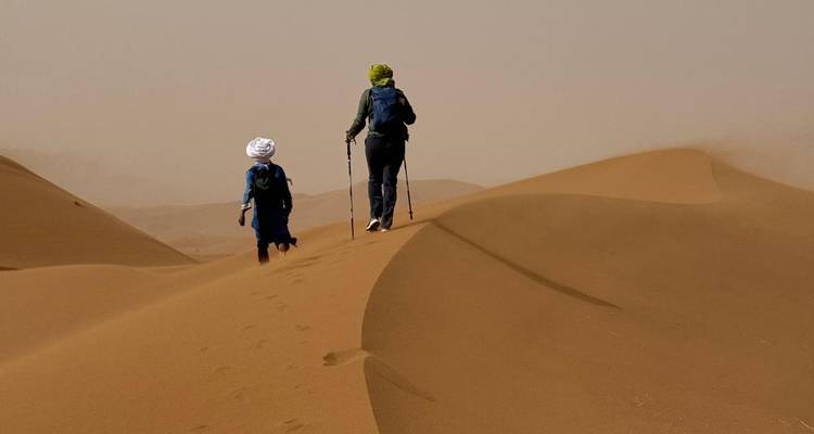 Dos excursionistas caminan por imponentes dunas de arena dorada bajo un cielo brumoso.