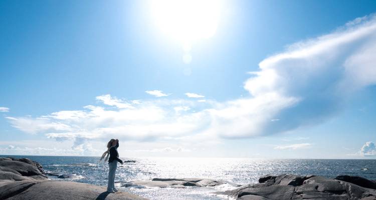 Mujer silueteada de pie en rocas costeras extendiendo los brazos hacia un sol brillante sobre un mar reluciente.