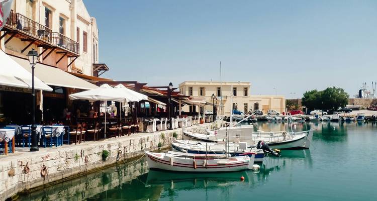Personas cenando junto al puerto con barcos atracados, posiblemente en Rethymnon, Grecia.
