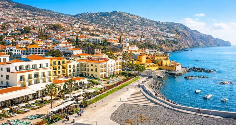 Une promenade ensoleillée et un port de plaisance font face au paysage urbain coloré de Funchal contre les collines verdoyantes qui s'élèvent.