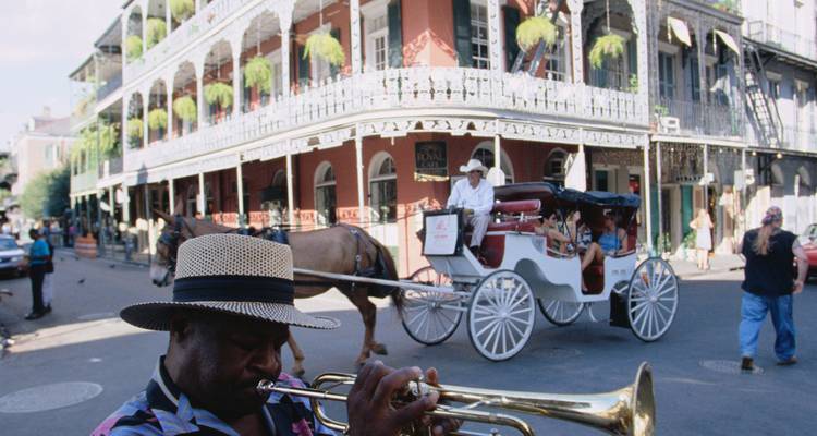 Un trompetista toca en el Barrio Francés de Nueva Orleans mientras un carruaje tirado por caballos pasa junto a balcones ornamentados.