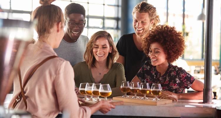 Groupe d'amis dans un bar de brasserie artisanale dégustant un plateau de bières servi par un barman.