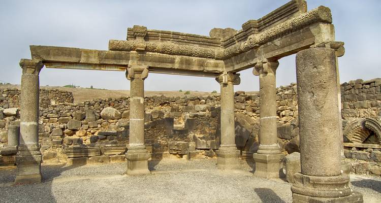 Ruins of a basalt stone synagogue with ornate lintel and four standing columns on a dry plateau.