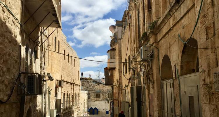 Sunlit narrow lane between high stone walls in Jerusalem’s Old City under blue sky.