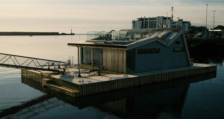 Modern zwevend sauna-huisje afgemeerd op rustige havenwateren onder zacht avondlicht.