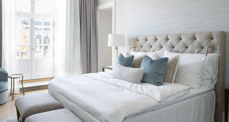 Licht en ruim hotelkamertje met gechaptonneerd hoofdeinde, witte lakens en pastelgekleurde kussens.