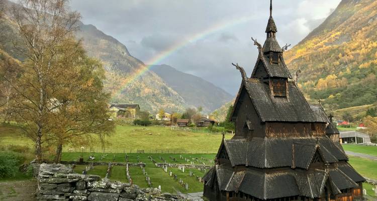 Mittelalterliche Stabkirche vor herbstlichen Bergen mit einem Regenbogen, der sich über dem Himmel spannt.