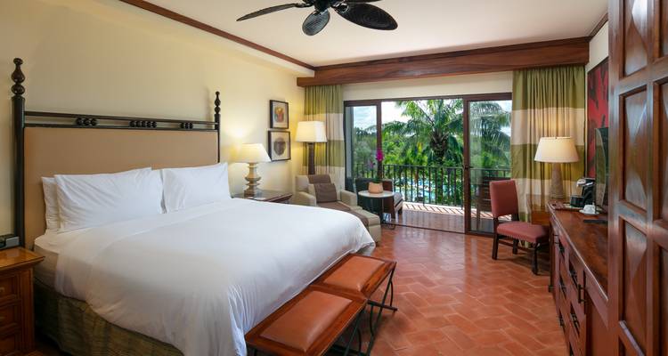 Chambre spacieuse du complexe hôtelier avec balcon offrant une vue sur les palmiers et la piscine, éclairage chaleureux et sol en terre cuite.
