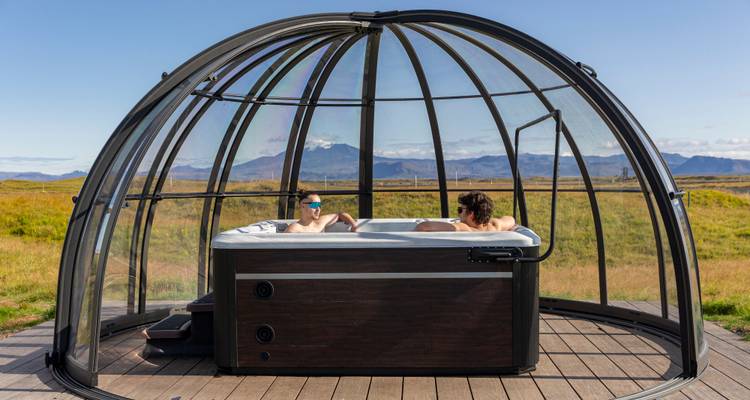 Un couple se détend dans un bain à remous extérieur surmonté d'un dôme en verre, profitant de la vue ensoleillée sur les montagnes.