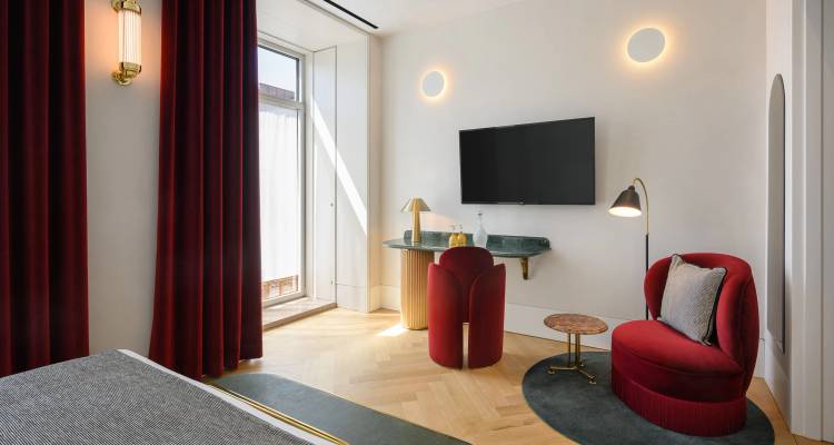 Habitación de hotel moderna y elegante con sillón de terciopelo rojo, escritorio pequeño, TV montada en la pared y una gran ventana que deja entrar mucha luz natural
