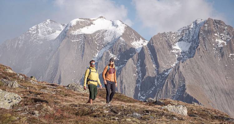 Dos excursionistas caminando por un sendero de montaña con picos nevados al fondo.