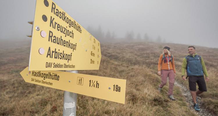 Excursionistas caminando junto a un letrero en un sendero montañoso con niebla.