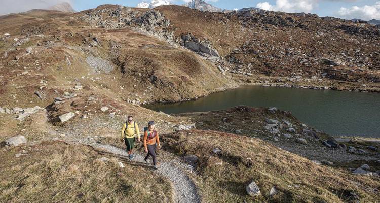 Dos excursionistas caminando por un sendero rocoso con un lago y montañas de fondo.