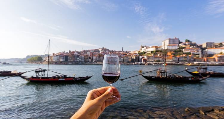 Hand hält ein Glas Portwein im Fokus mit Portos Ribeira-Viertel und traditionellen Rabelo-Booten am Douro-Fluss.