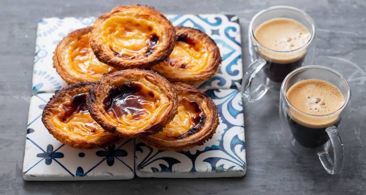 Nahaufnahme von traditionellen portugiesischen Pastéis de Nata auf dekorativen Fliesen mit Espresso-Shots daneben.