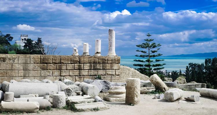 Colonnes de marbre en ruine et blocs de pierre de Carthage surplombant une mer turquoise et des collines.