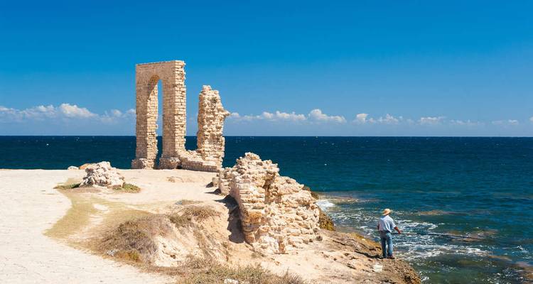 Un pêcheur solitaire se tient près des ruines d'une arche de pierre ancienne sur un promontoire rocheux méditerranéen.