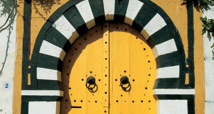 Porte tunisienne traditionnelle jaune vif encadrée par un arc en fer à cheval noir et blanc.