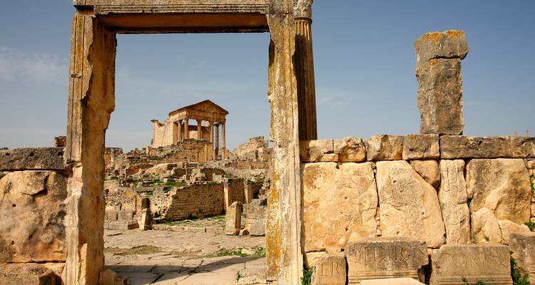 Une porte de pierre ancienne encadre les imposantes ruines du Capitole romain au milieu du site archéologique de Dougga sous un ciel dégagé.