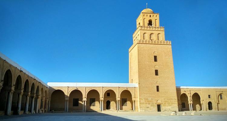 La cour ensoleillée de la Grande Mosquée de Kairouan avec son haut minaret carré se dressant contre un ciel bleu profond.