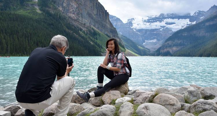 Voyageur posant sur les rochers du bord du lac tandis que son compagnon le photographie, avec le lac Louise turquoise et les glaciers au-delà