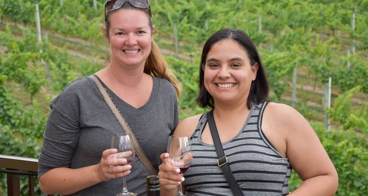 Deux femmes souriantes avec des verres de vin à la main devant un décor de rangées de vignes luxuriantes