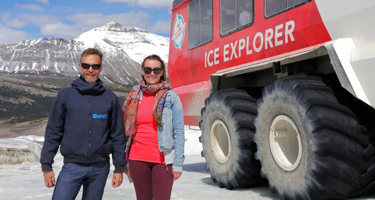 Voyageurs posant à côté du véhicule rouge Ice Explorer sur un glacier enneigé avec des pics rocheux en arrière-plan