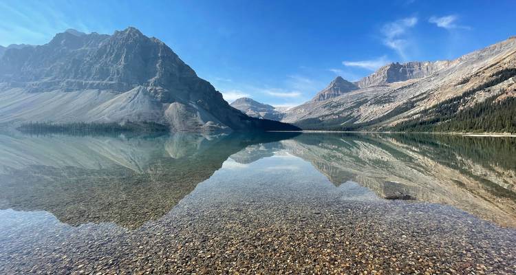 Lac alpin semblable à un miroir reflétant des pics escarpés et un ciel sans nuages, rivage de galets au premier plan