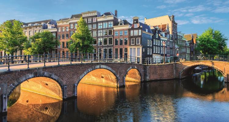 Icónico puente de ladrillo arqueado sobre un canal reflectante bordeado de casas adosadas altas y estrechas de Ámsterdam bajo una cálida luz vespertina.