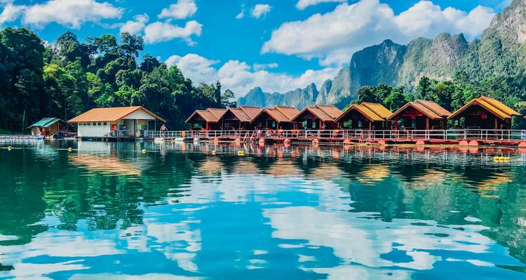 Schwimmendes Holzbungalow-Resort auf türkisem See, umgeben von üppigem Dschungel und Kalksteinfelsen unter hellem Himmel.