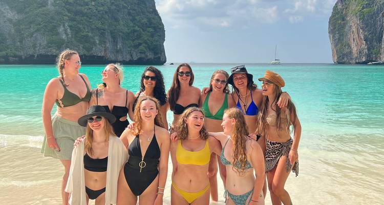Lachende Gruppe von Frauen in Badebekleidung, die an einem tropischen weißen Sandstrand mit smaragdgrünem Wasser und Kalksteinfelsen posieren.