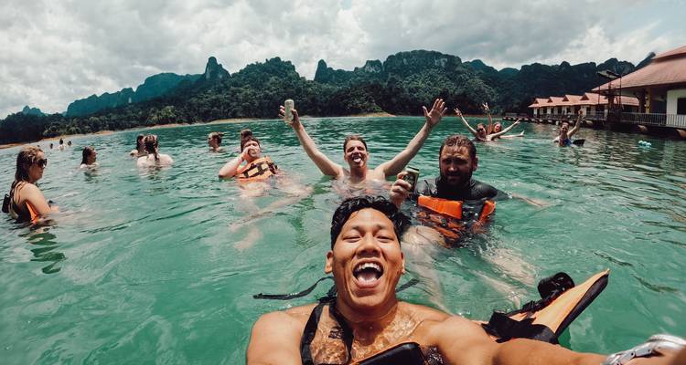 Selfie von fröhlichen Reisenden, die schwimmen und mit Getränken in einem jadegrünen See jubeln, umgeben von bewaldeten Gipfeln.