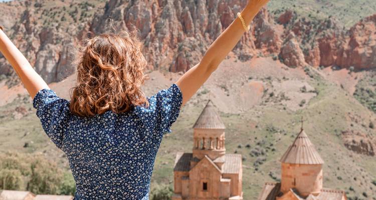 Een vrouw steekt haar armen omhoog van vreugde terwijl ze uitkijkt over de stenen kerken van Noravank tegen rode kliffen.