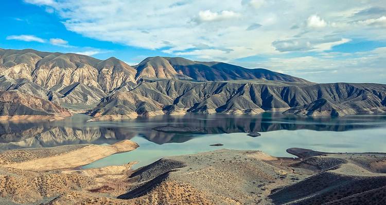 Een uitgestrekt bergmeer weerkaatst ruige bruine bergkammen en een lappendeken van kustlijn onder verspreide wolken.