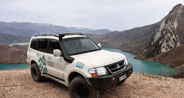 Véhicule tout-terrain 4x4 garé sur un point de vue rocheux surplombant un lac albanais turquoise et des montagnes.
