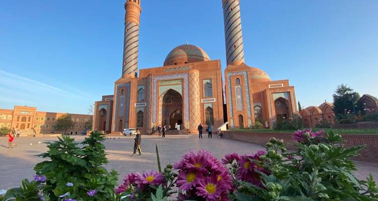 Grande mosquée en brique avec des minarets jumeaux rayés et un dôme central encadrée par des fleurs violettes en fleurs au premier plan sous un ciel bleu dégagé