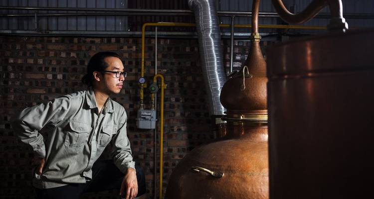 Un jeune distillateur étudie un grand alambic en cuivre à l'intérieur d'un atelier aux murs en brique.