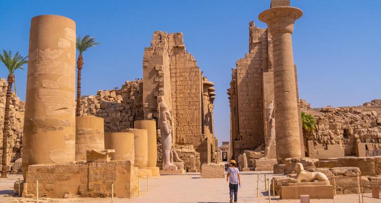 Persona caminando frente a ruinas del antiguo Egipto con estatuas and columnas bajo un cielo azul despejado.