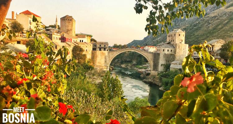 Vue pittoresque de l'emblématique pont en arc Stari Most enjambant une rivière verte, encadré par les maisons colorées du village et de vives fleurs dans la douce lumière du soir.