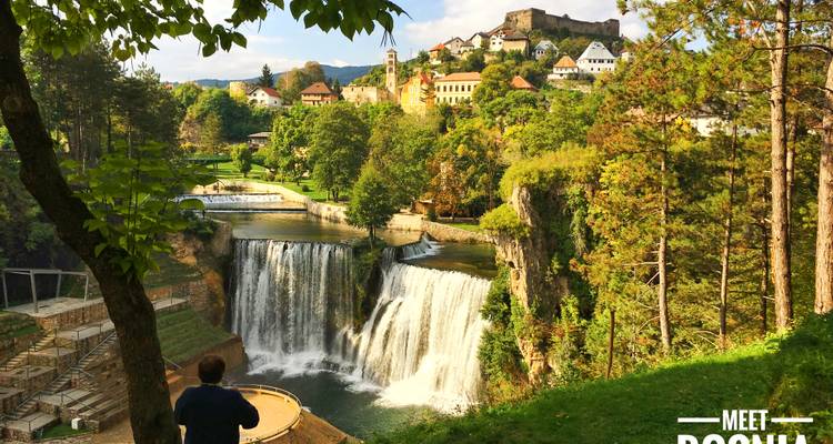 Point de vue surélevé sur une cascade spectaculaire à plusieurs niveaux avec une ville historique au-dessus et un visiteur admirant la scène depuis une plateforme.
