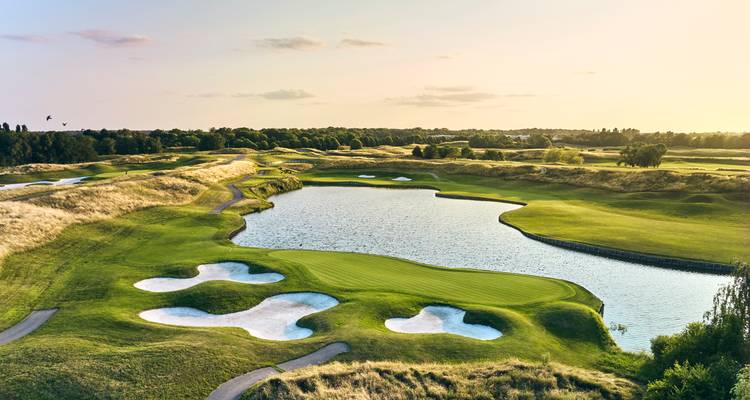 Parcours de golf ensoleillé avec un grand étang réfléchissant et des bunkers aux formes artistiques.
