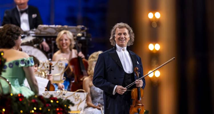 Orchesteraufführung mit einem lächelnden Geiger in formeller Kleidung, der im Zentrum der Bühne unter warmen Scheinwerfern steht.